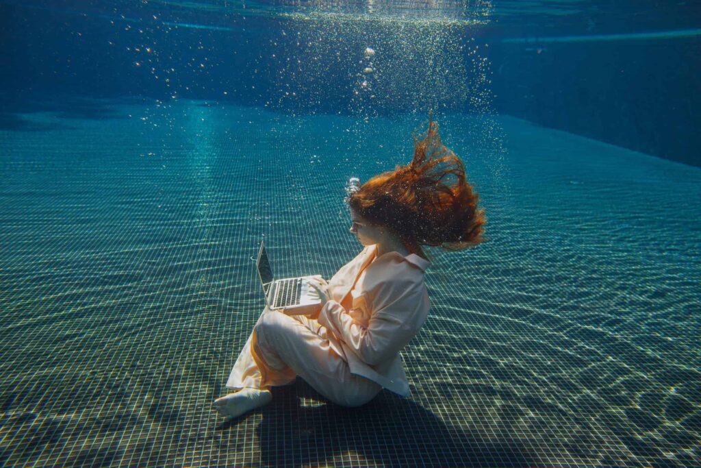 A woman in a pale suit sits on the floor of a swimming pool, underwater, typing on a laptop.