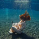 A woman in a pale suit sits on the floor of a swimming pool, underwater, typing on a laptop.