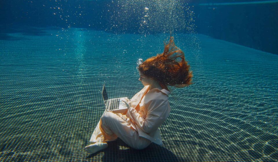 A woman in a pale suit sits on the floor of a swimming pool, underwater, typing on a laptop.