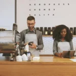 Two café staff behind the counter, one drying a cup, one at a screen