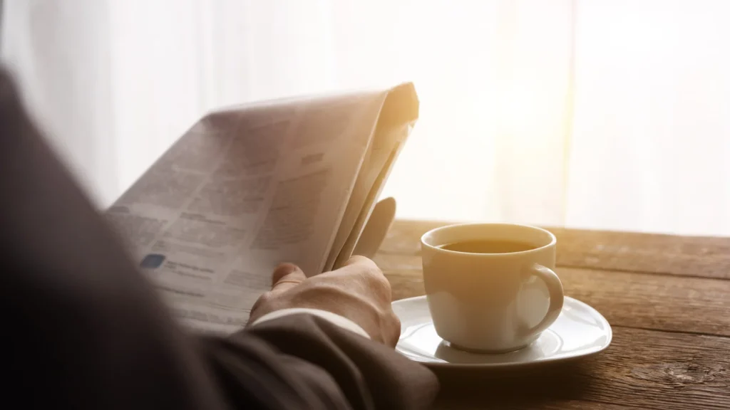 A man reading a newspaper in morning light, a cup of coffee on the table