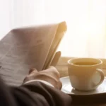A man reading a newspaper in morning light, a cup of coffee on the table