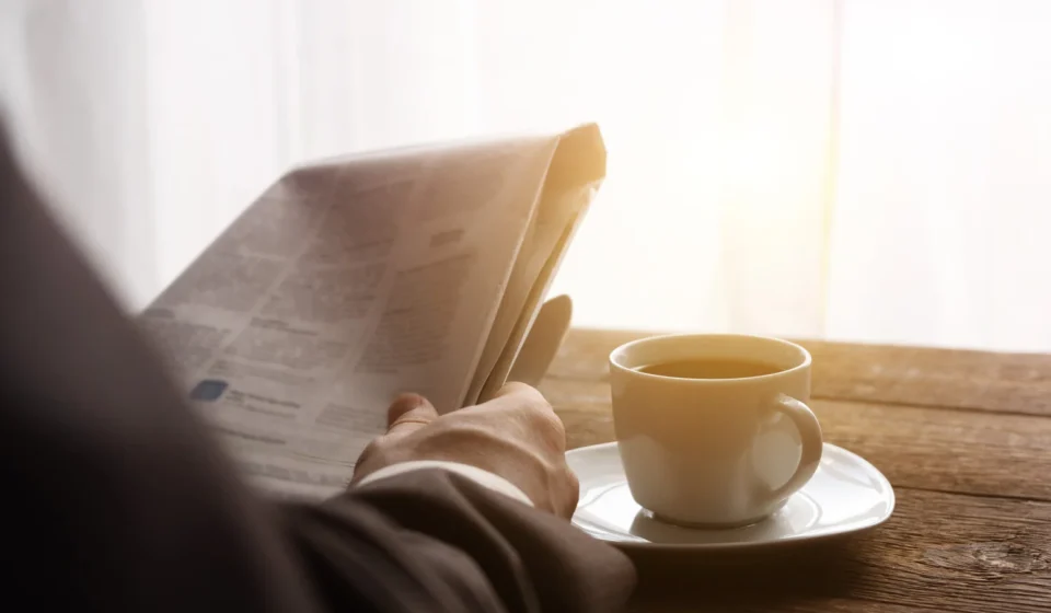 A man reading a newspaper in morning light, a cup of coffee on the table