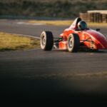 A red open-wheel single-seater race car taking a corner on a race track in low evening light, its driver in a dark closed helmet.