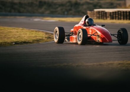 A red open-wheel single-seater race car taking a corner on a race track in low evening light, its driver in a dark closed helmet.