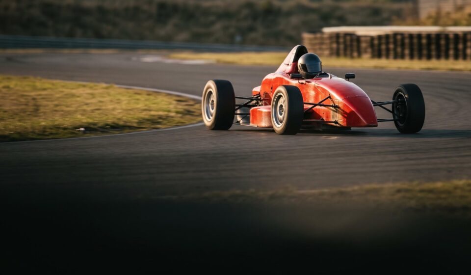 A red open-wheel single-seater race car taking a corner on a race track in low evening light, its driver in a dark closed helmet.