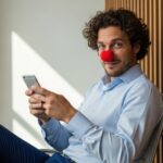 A man in a light blue business shirt sits with his phone and smiles at the camera, wearing a red clown nose.