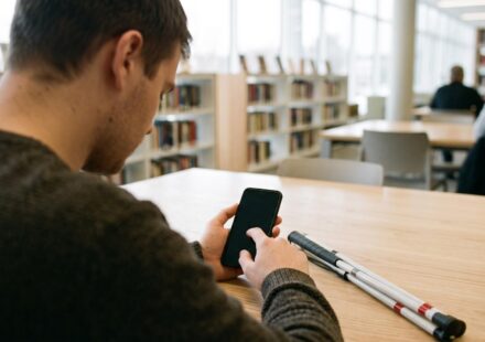 A young blind man at a library table swipes through a phone with its screen switched off, listening to it read aloud, a folded white cane beside his arm.