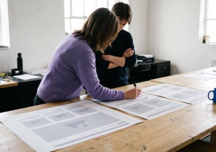 An agency owner circles a flaw on a printed proof of a web page laid out at three sizes, while a colleague watches.