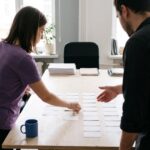 An agency owner briefs a project manager, who lays blank task cards in columns on a large table.