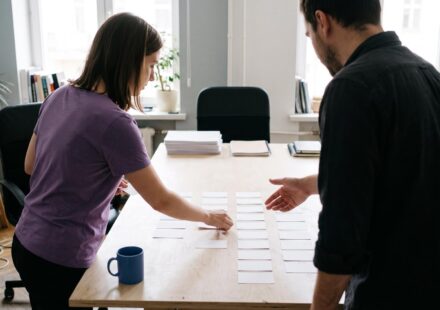 An agency owner briefs a project manager, who lays blank task cards in columns on a large table.