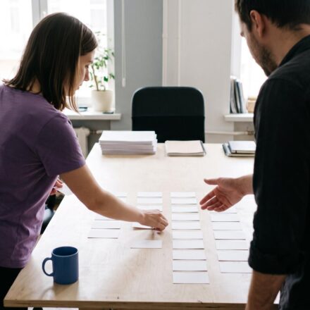 An agency owner briefs a project manager, who lays blank task cards in columns on a large table.