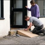 A carpenter kneels at a restaurant doorway and screws a pale oak ramp over the stone step while the owner, in a violet sweater, holds the door open.