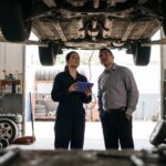 A mechanic holding a diagnostic tablet and a car owner stand under a car raised on a lift, both looking up at its underbody.