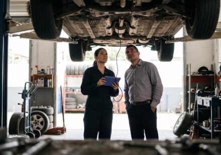 A mechanic holding a diagnostic tablet and a car owner stand under a car raised on a lift, both looking up at its underbody.