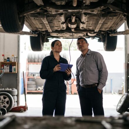 A mechanic holding a diagnostic tablet and a car owner stand under a car raised on a lift, both looking up at its underbody.