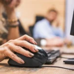 A woman's hand on a black computer mouse, the scroll wheel visible beside her index finger, at an office desk with a monitor