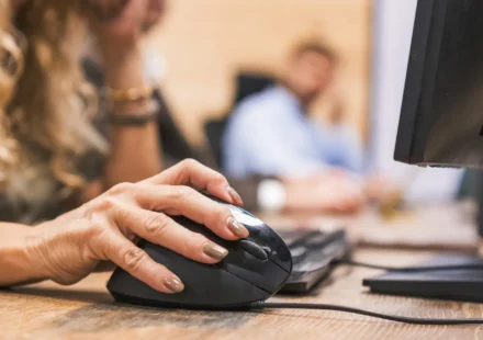 A woman's hand on a black computer mouse, the scroll wheel visible beside her index finger, at an office desk with a monitor