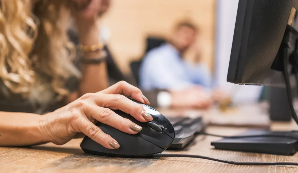 A woman's hand on a black computer mouse, the scroll wheel visible beside her index finger, at an office desk with a monitor
