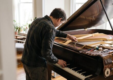 A piano tuner leaning into an open grand piano, turning a tuning pin with one hand and pressing a key with the other, his tool roll laid on the case.