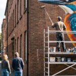 A painter on a scaffold tower, seen from behind, rolling paint onto a large mural of a kingfisher on a brick gable wall, while two passers-by on the pavement stop to look up at it.