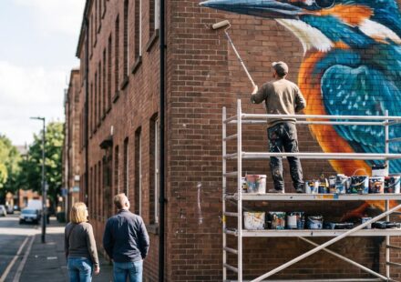 A painter on a scaffold tower, seen from behind, rolling paint onto a large mural of a kingfisher on a brick gable wall, while two passers-by on the pavement stop to look up at it.
