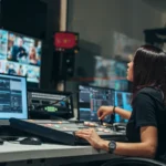 Una operadora en una sala de control de televisión, frente a una mesa de mezcla y varios monitores