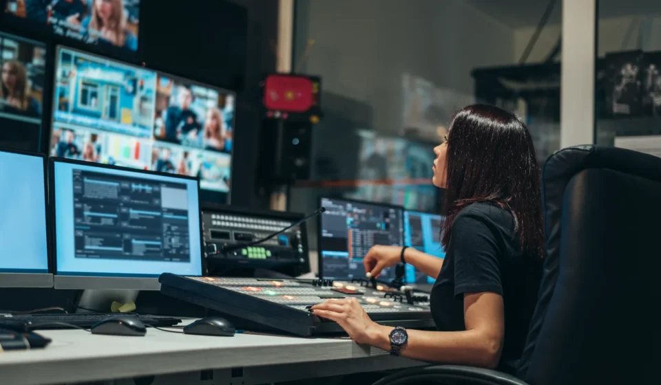 Una operadora en una sala de control de televisión, frente a una mesa de mezcla y varios monitores