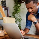 A bakery owner leaning on his counter, reading a laptop beside fresh bread