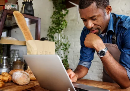 A bakery owner leaning on his counter, reading a laptop beside fresh bread