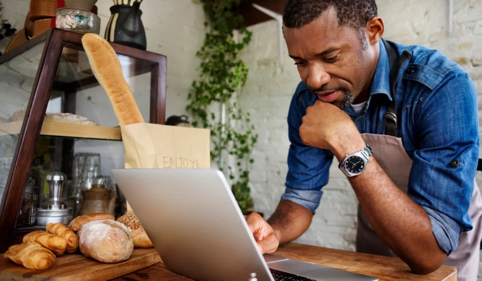 A bakery owner leaning on his counter, reading a laptop beside fresh bread
