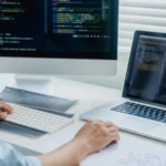 A developer working across a desktop monitor and a laptop, both showing code