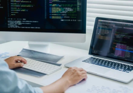 A developer working across a desktop monitor and a laptop, both showing code