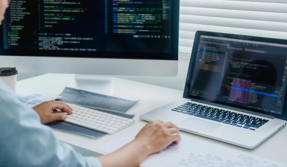 A developer working across a desktop monitor and a laptop, both showing code