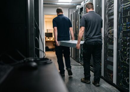 Two technicians, seen from behind, carrying a rack server down an aisle between server cabinets in a server room.