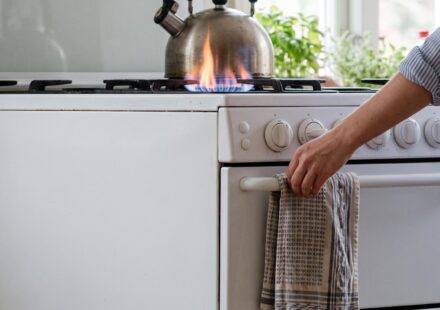 A forearm in a rolled striped sleeve resting across the knobs of a white gas cooker while the hand hangs a tea towel on the oven door, and the flame under the kettle flares high.