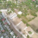 Vista aérea de un terreno despejado con una calle nueva y casas recién construidas, junto a un barrio de casas y árboles