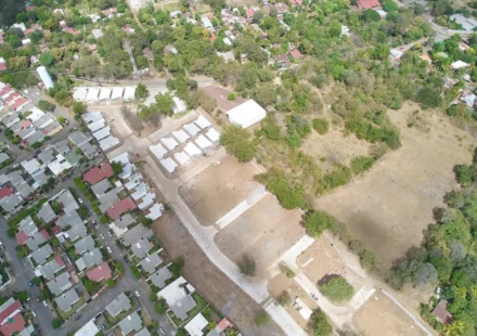 Vista aérea de un terreno despejado con una calle nueva y casas recién construidas, junto a un barrio de casas y árboles