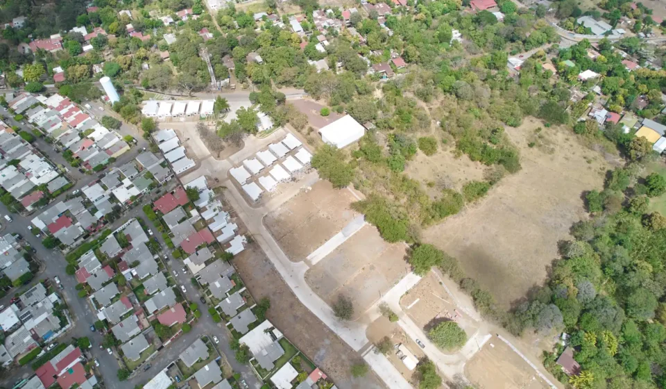 Vista aérea de un terreno despejado con una calle nueva y casas recién construidas, junto a un barrio de casas y árboles