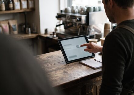 A café owner in an apron, seen over the shoulder, tapping a toggle switch on a tablet that stands on a wooden counter, with an espresso machine behind.