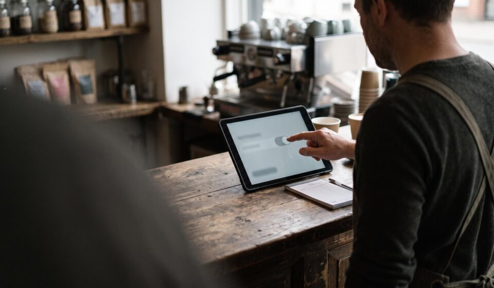 A café owner in an apron, seen over the shoulder, tapping a toggle switch on a tablet that stands on a wooden counter, with an espresso machine behind.