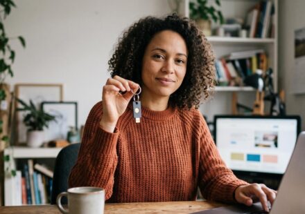 A woman with curly hair in a rust-coloured knit sitting at a desk, holding up a small security key on a ring and looking at the camera, her other hand on the keyboard of an open laptop.