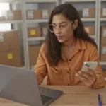 A shop owner at a laptop, phone in hand, in a stockroom of labelled parcels