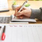 A hand ticking items off a written checklist beside an open laptop