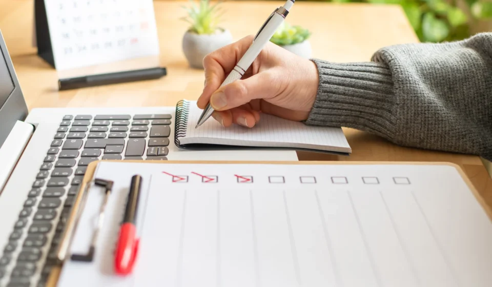 A hand ticking items off a written checklist beside an open laptop