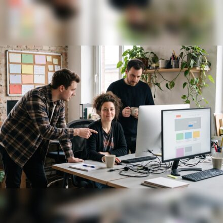 Three colleagues at a shared desk in a small studio: a woman seated in the middle looks at the camera, a man in a checked shirt leans in and points at a monitor showing a page layout in coloured blocks, and a third stands behind holding a mug.