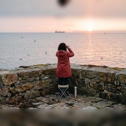 A person in a red coat, seen from behind, looking through binoculars over a stone harbour wall at a ship on the horizon at sunrise.