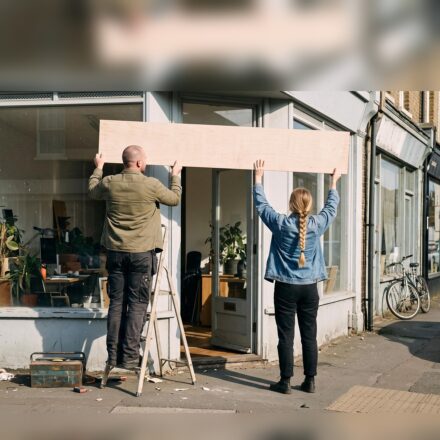 Two people, seen from behind, lifting a long blank wooden signboard into place above the door of a small studio: a man in an olive overshirt on a stepladder and a woman with a long blonde plait in a denim jacket on the pavement.